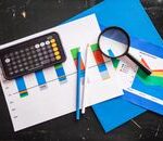 Top view of financial charts with a smartphone calculator, magnifying glass, and pencils on a desk.